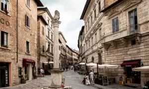 Strada di Montepulciano con un leone di pietra, negozi e gente. Un cartello pubblicizza degustazioni di vino.