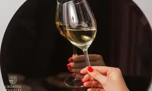 Un brindisi con i vini croati. Due calici di vino bianco dorato, tenuti da mani con smalto rosso, riflettono un'atmosfera elegante.