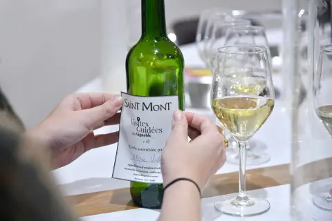 Mani che applicano un'etichetta a una bottiglia di vino bianco Saint Mont, con bicchieri pieni in primo piano. Un assaggio del **madiran**?