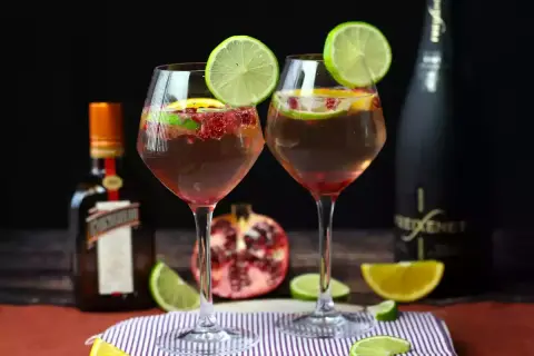 Due calici di sangria con prosecco, guarniti con lime e melograno, accanto a bottiglie di Cointreau e Freixenet.