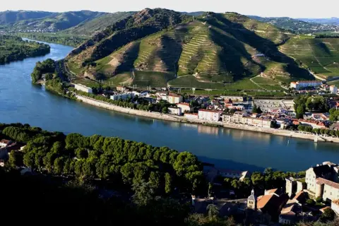 Villaggi pittoreschi lungo il Rodano, con vigneti terrazzati che salgono sui pendii verdi. Un fiume serpeggia tra i borghi, evocando l'atmosfera dei Cotes du Rhone Villages.