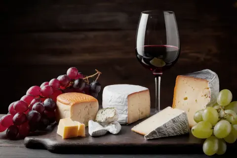 Assortment of cheeses, grapes, and a glass of red wine on a dark wooden board.