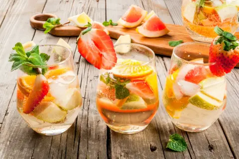 Tre bicchieri di sangria con vino bianco, fragole, arance, mele e menta su un tavolo di legno.