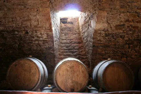 Cantine in pietra con botti di legno, un raggio di luce illumina la scala. Un'atmosfera che evoca il **Rodano, Francia**, e i suoi vini pregiati.