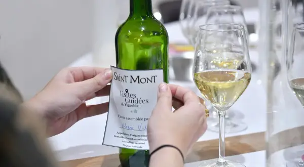 Mani che applicano un'etichetta a una bottiglia di vino bianco Saint Mont, con bicchieri pieni in primo piano. Un assaggio del **madiran**?