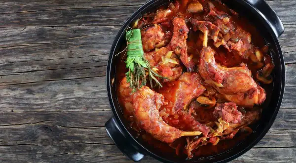 Un saporito coniglio al vino rosso, cotto lentamente con erbe aromatiche, in una casseruola nera su un tavolo di legno rustico.