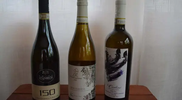 Tre bottiglie di vino bianco, tra cui un Lison Classico di Pramaggiore, su un tavolo di legno.