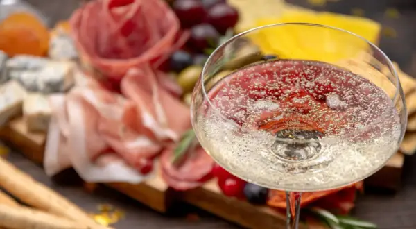 Un bicchiere di spumanti italiani famosi, con un tagliere di salumi, formaggi e uva sullo sfondo.