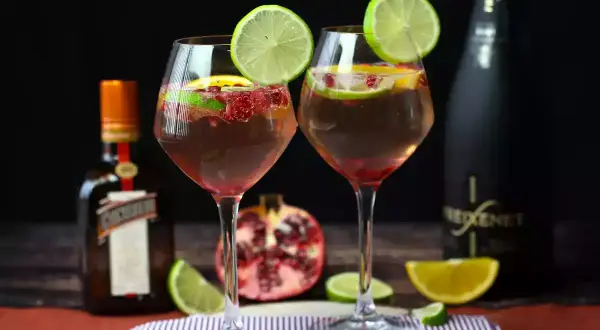 Due calici di sangria con prosecco, guarniti con lime e melograno, accanto a bottiglie di Cointreau e Freixenet.