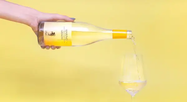 Versando vino bianco Marino Superiore, un'eccellenza tra i vini romani, in un calice su sfondo giallo.