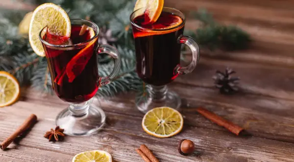 Due bicchieri di vino per vin brulé, decorati con fette d'arancia e cannella, su un tavolo di legno con rami di pino.