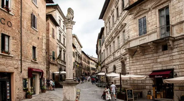 Strada di Montepulciano con un leone di pietra, negozi e gente. Un cartello pubblicizza degustazioni di vino.