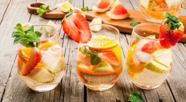 Tre bicchieri di sangria con vino bianco, fragole, arance, mele e menta su un tavolo di legno.