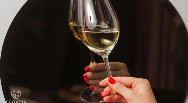 Un brindisi con i vini croati. Due calici di vino bianco dorato, tenuti da mani con smalto rosso, riflettono un'atmosfera elegante.