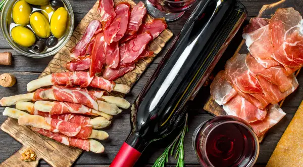 Un bicchiere e una bottiglia di vino rosso bordeaux, accompagnati da salumi, grissini, olive e noci, creano un'atmosfera conviviale.
