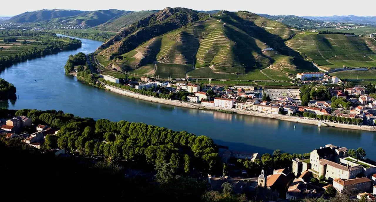Villaggi pittoreschi lungo il Rodano, con vigneti terrazzati che salgono sui pendii verdi. Un fiume serpeggia tra i borghi, evocando l'atmosfera dei Cotes du Rhone Villages.