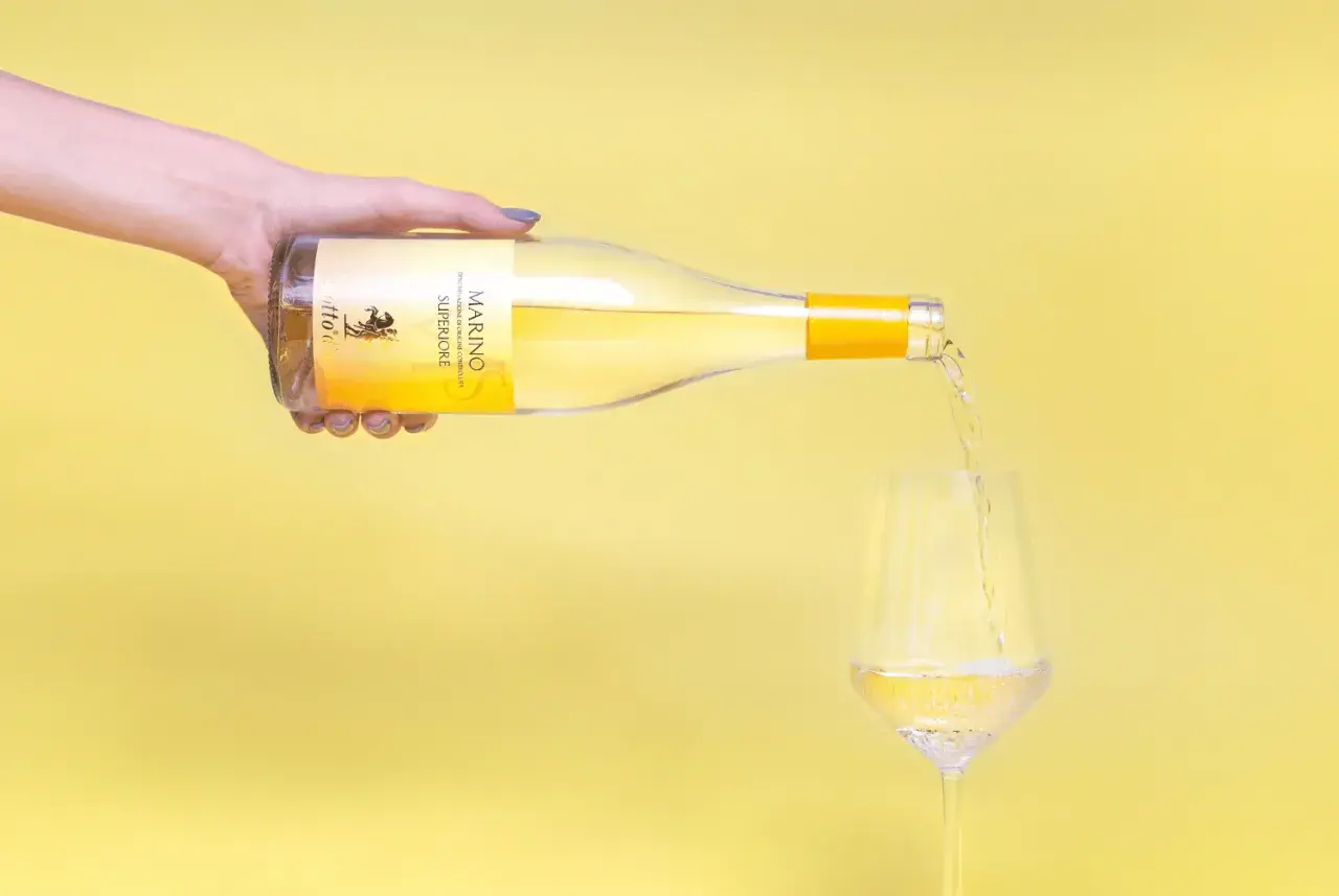 Versando vino bianco Marino Superiore, un'eccellenza tra i vini romani, in un calice su sfondo giallo.
