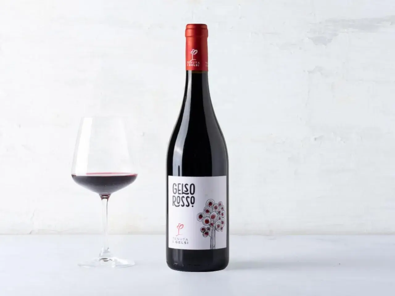 A bottle of red wine and a glass of red wine on a white surface.