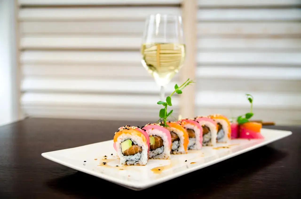 Rotolino di sushi colorato su un piatto bianco, con un bicchiere di vino da abbinare al sushi sullo sfondo.