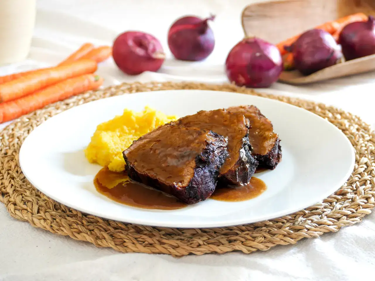 Arrosto succulento in salsa, accompagnato da polenta. Un classico tra le ricette con il vino rosso, con cipolle e carote sullo sfondo.