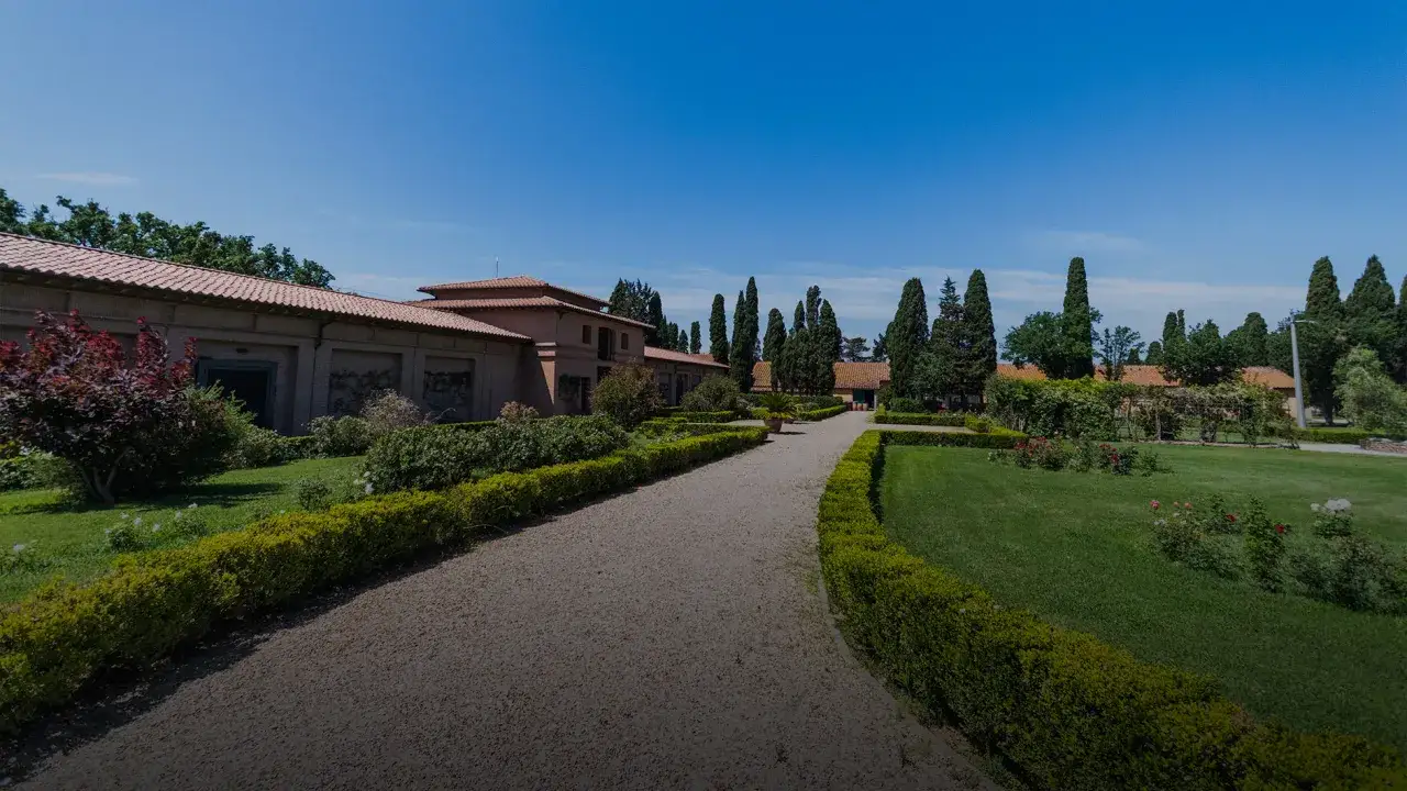 Giardini curati e un viale ghiaioso conducono a un edificio con tetto in tegole rosse, evocando l'atmosfera del Sassicaia origine.