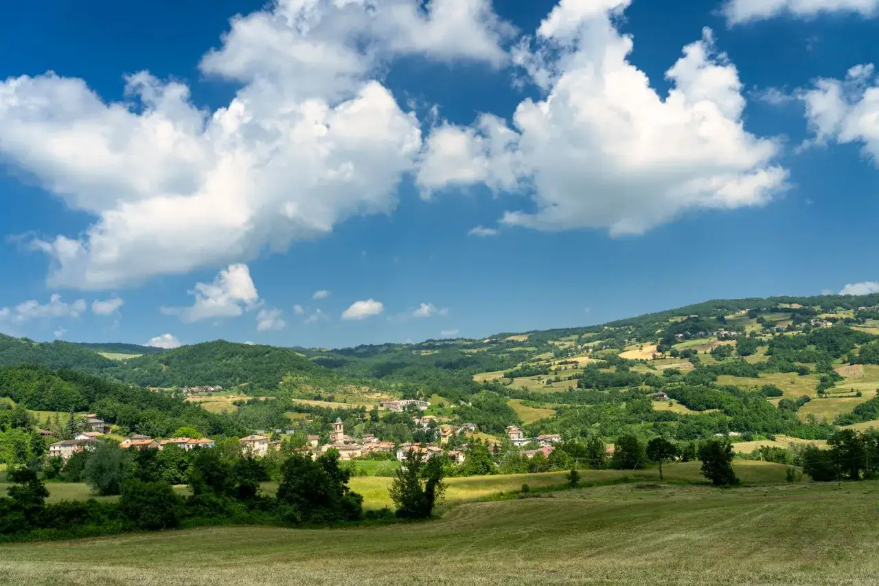 Un borgo incantevole tra i verdi colli piacentini, sotto un cielo azzurro punteggiato di nuvole bianche.