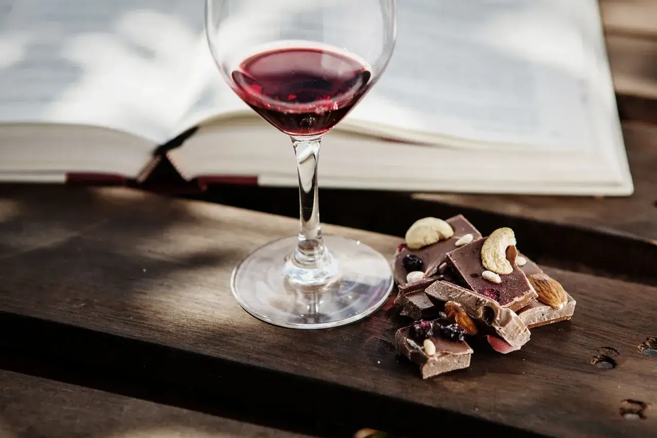 Un bicchiere di vino rosso accanto a pezzi di cioccolato con frutta secca e noci, con un libro aperto sullo sfondo.