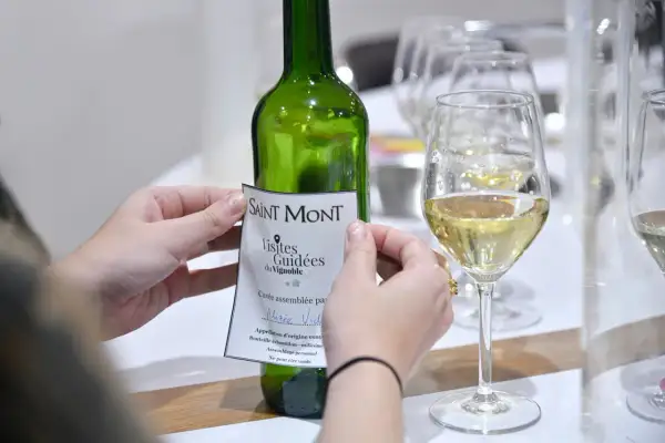 Mani che applicano un'etichetta a una bottiglia di vino bianco Saint Mont, con bicchieri pieni in primo piano. Un assaggio del **madiran**?