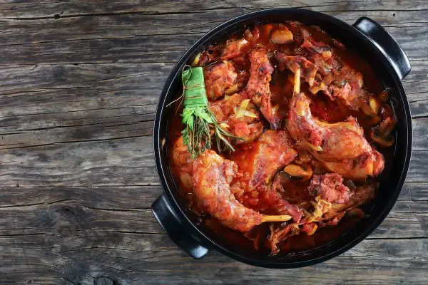 Un saporito coniglio al vino rosso, cotto lentamente con erbe aromatiche, in una casseruola nera su un tavolo di legno rustico.