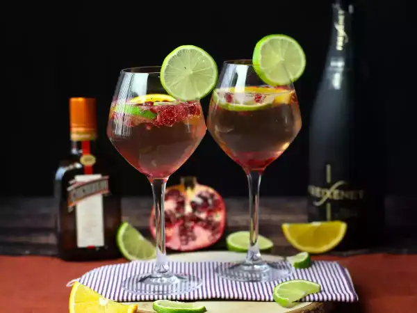 Due calici di sangria con prosecco, guarniti con lime e melograno, accanto a bottiglie di Cointreau e Freixenet.