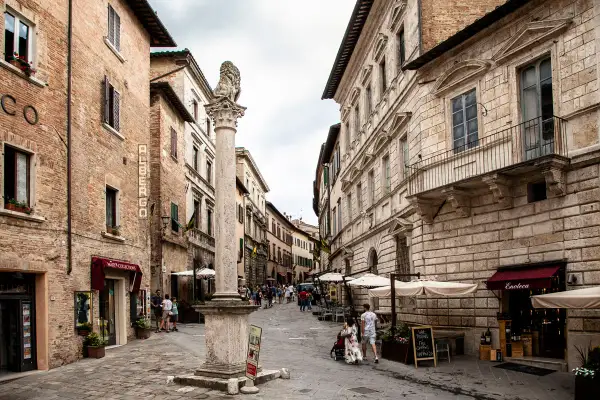 Strada di Montepulciano con un leone di pietra, negozi e gente. Un cartello pubblicizza degustazioni di vino.
