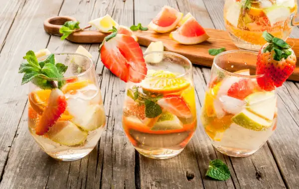 Tre bicchieri di sangria con vino bianco, fragole, arance, mele e menta su un tavolo di legno.