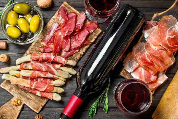 Un bicchiere e una bottiglia di vino rosso bordeaux, accompagnati da salumi, grissini, olive e noci, creano un'atmosfera conviviale.